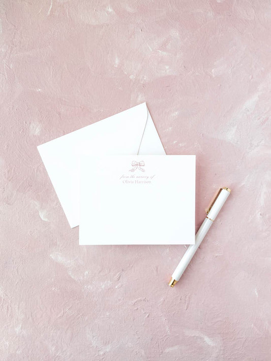 Elegant flat notecard with a delicate pink bow design and personalized text, paired with a white envelope and gold-accented pen on a soft pink background.