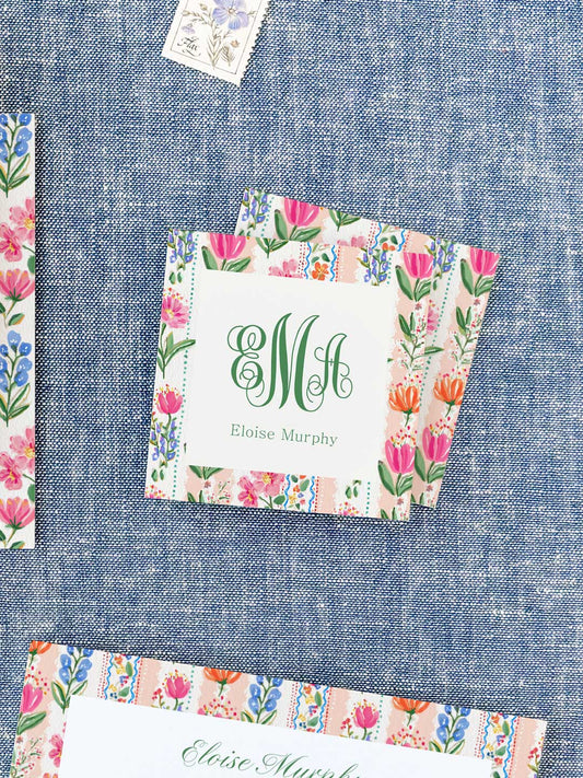 Square calling cards with colorful floral borders and elegant green monogram and name on a textured blue background
