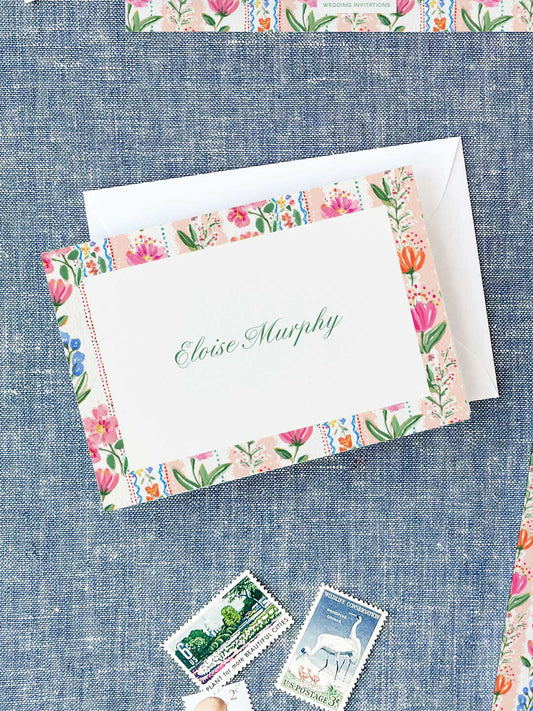 Folded notecard with colorful floral border and elegant green script name on textured blue fabric background.