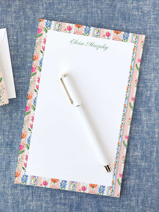 Notepad with colorful floral border and personalized name at the top, next to a white pen on blue fabric.