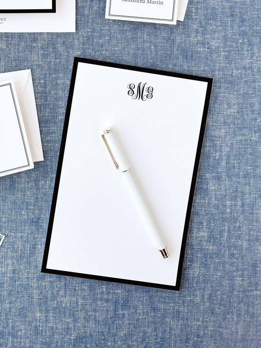 Personalized notepad featuring a classic black border and elegant monogram, displayed with a white pen on textured fabric background.