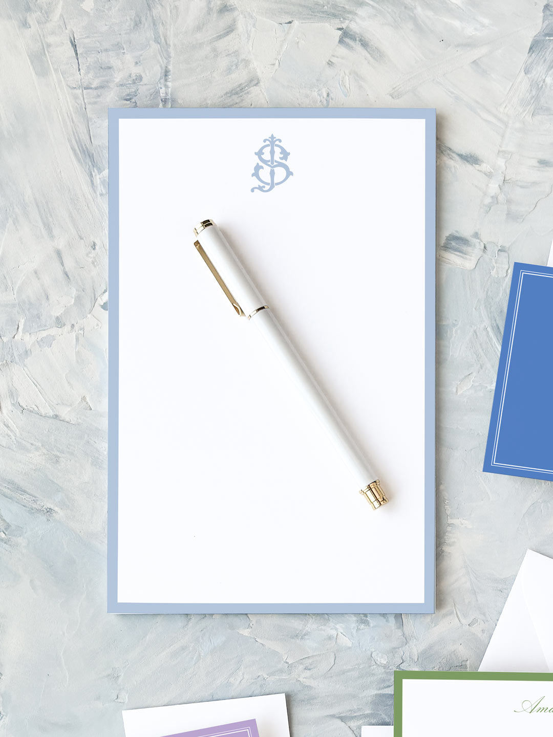 White pen resting on a pale blue-bordered notepad with a subtle decorative monogram on textured gray surface.