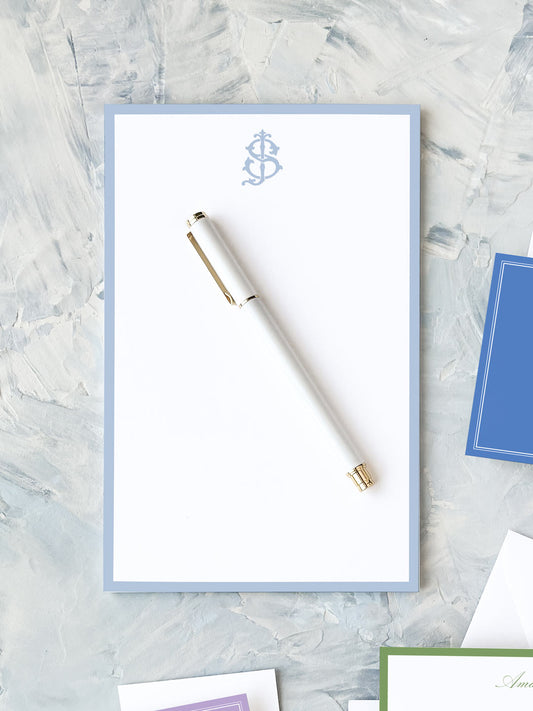White pen resting on a pale blue-bordered notepad with a subtle decorative monogram on textured gray surface.