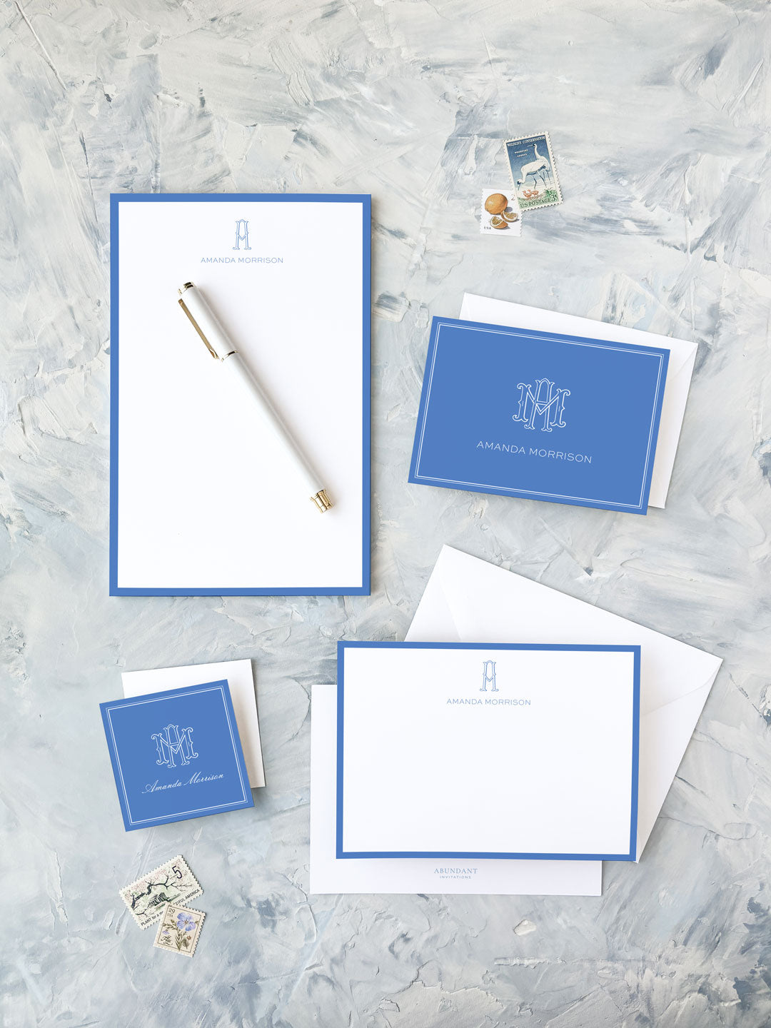 Elegant personalized stationery set in cobalt blue featuring notepads, folded cards, envelopes, and a white pen on a textured light gray surface.