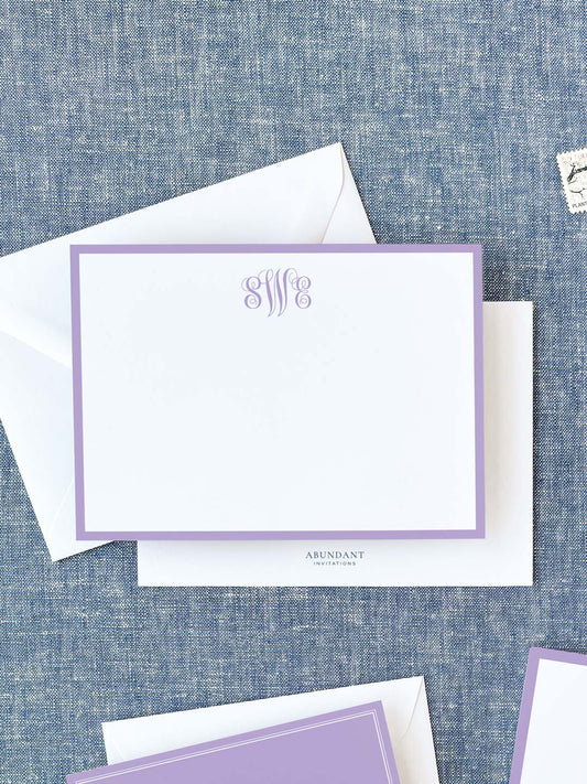 Elegant white flat notecard with a lavender border and monogram, paired with matching envelope on textured fabric background.