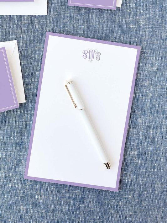 Notepad with a soft lavender border and elegant monogram lettering at the top, accompanied by a sleek white pen on a textured blue surface.