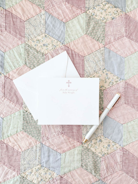 White notecard with a delicate pink cross and subtle text, paired with a white envelope and pen, displayed on a pastel-colored quilt background.
