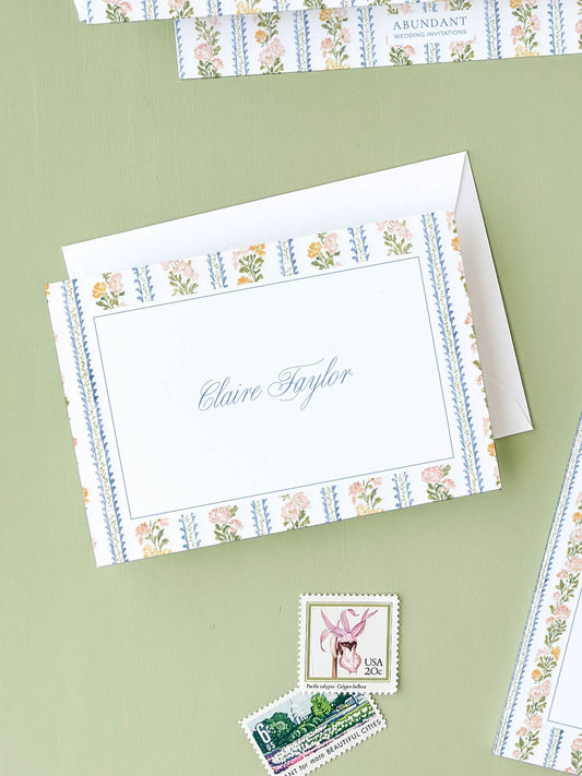 Folded notecard with floral and stripe border design resting on green surface with vintage postage stamps nearby