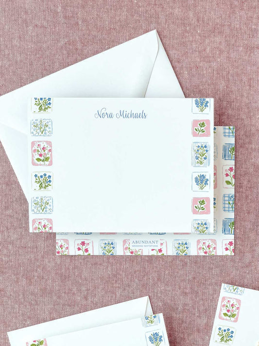 Personalized flat notecard with delicate floral tile border and matching envelope on a textured surface.