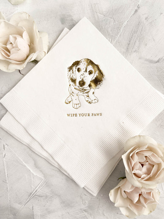White cocktail napkin with a detailed dog illustration and the phrase "wipe your paws" surrounded by cream roses on a textured surface.