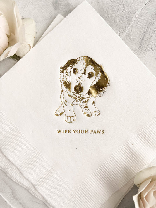 White napkin with gold foil custom dog illustration and "Wipe Your Paws" text in elegant script style