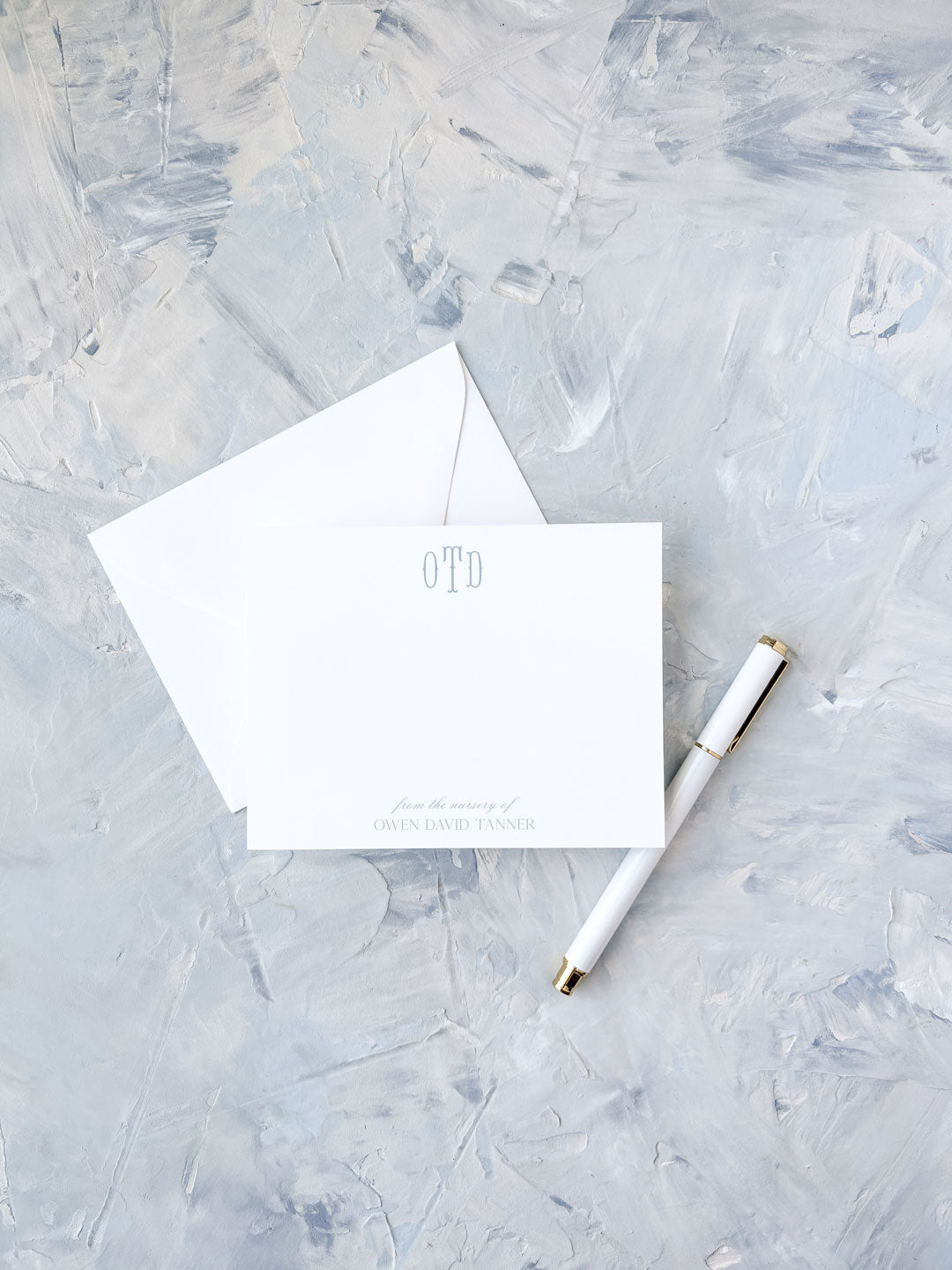 Elegant flat personalized stationery featuring a subtle blue monogram and matching envelope, styled with a white and gold pen on a textured light background.