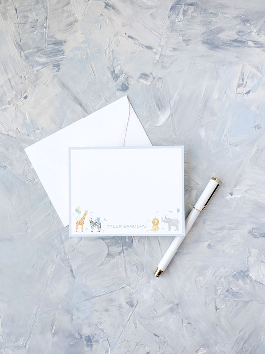 Personalized flat notecard with playful animal illustrations and light blue border, accompanied by a white envelope and elegant pen on textured background.