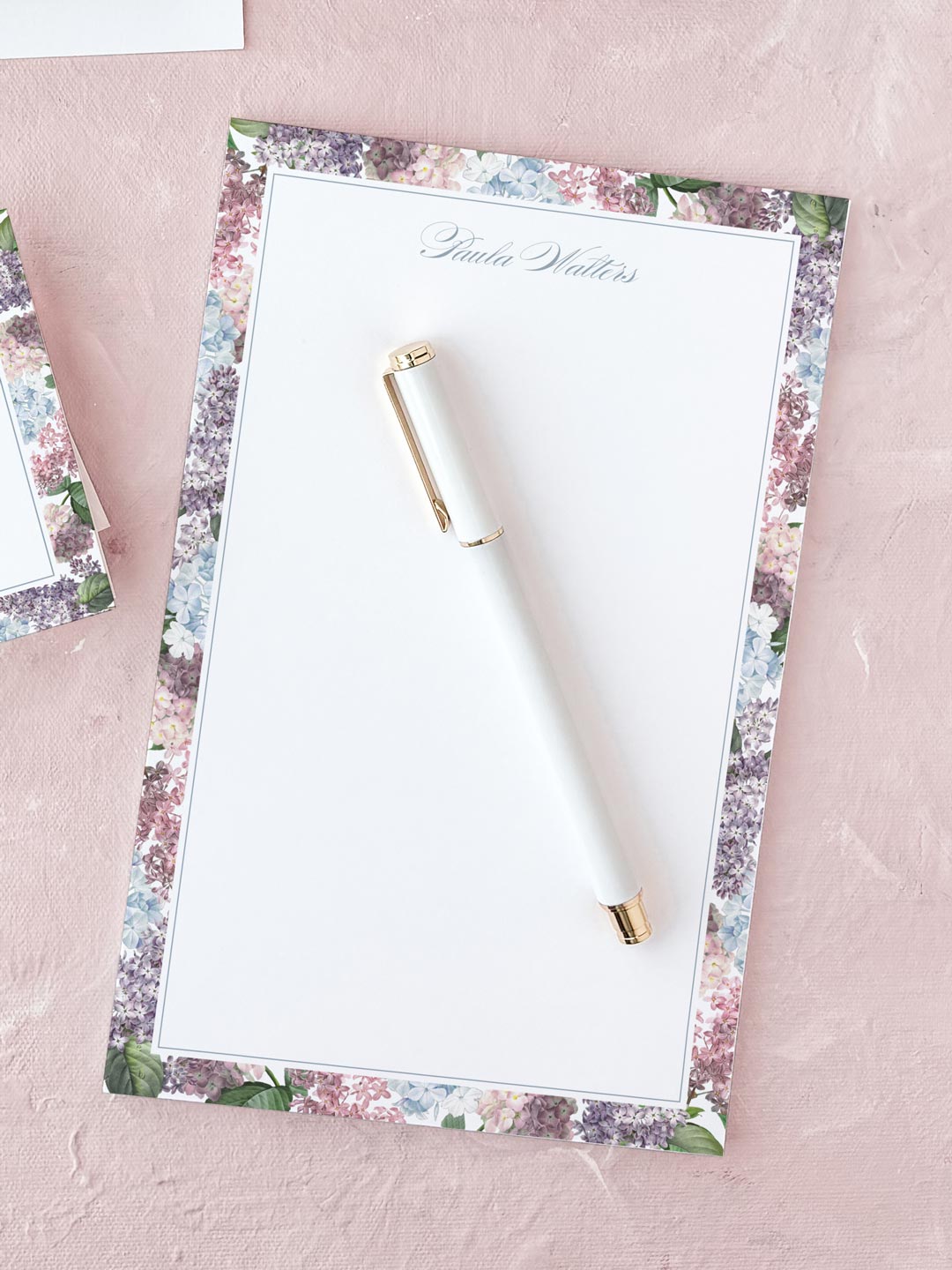Notepad with delicate floral border and elegant script, placed on soft pink fabric with white pen on top.