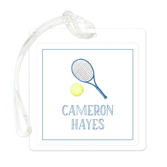 Clear rectangular bag tag featuring a blue tennis racket and yellow tennis ball illustration above the name Cameron Hayes in blue text, attached to a transparent strap.