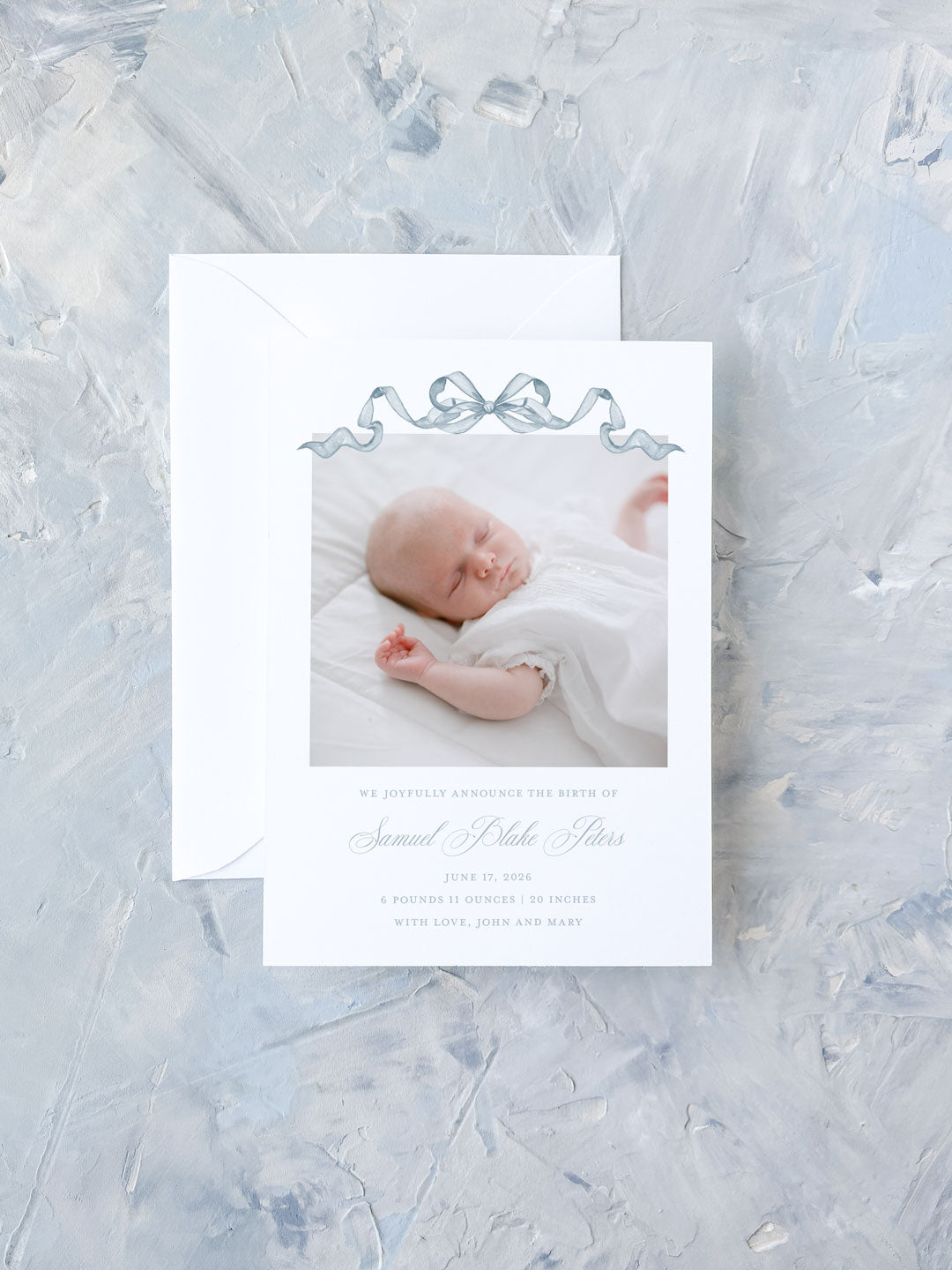 Birth announcement card featuring a sleeping baby photo bordered by a delicate blue watercolor bow, displayed with a white envelope on a textured pale background.