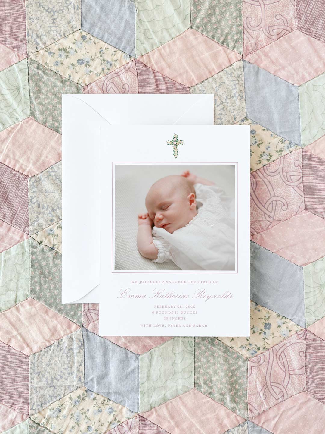 Birth announcement card featuring a soft watercolor floral cross and a photo of a sleeping newborn on a pastel patchwork quilt background.