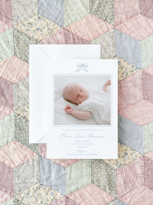 Birth announcement card with a soft blue bow illustration, featuring a photo of a sleeping newborn on a pastel patchwork quilt background.