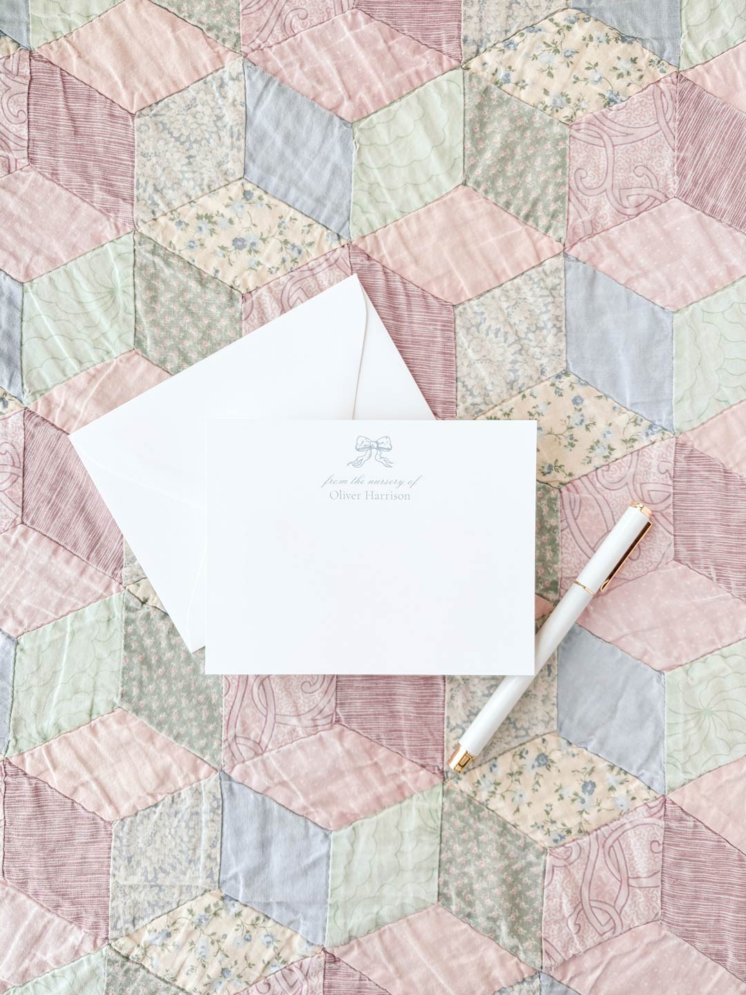 White flat notecard with a delicate blue bow design and personalized text, displayed with an envelope and pen on a pastel patchwork quilt background.