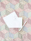 White flat notecard with a delicate blue bow design and personalized text, displayed with an envelope and pen on a pastel patchwork quilt background.