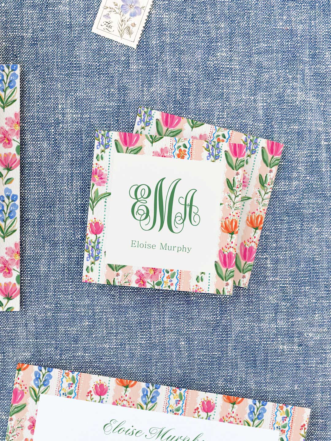 Square calling cards with colorful floral borders and elegant green monogram and name on a textured blue background
