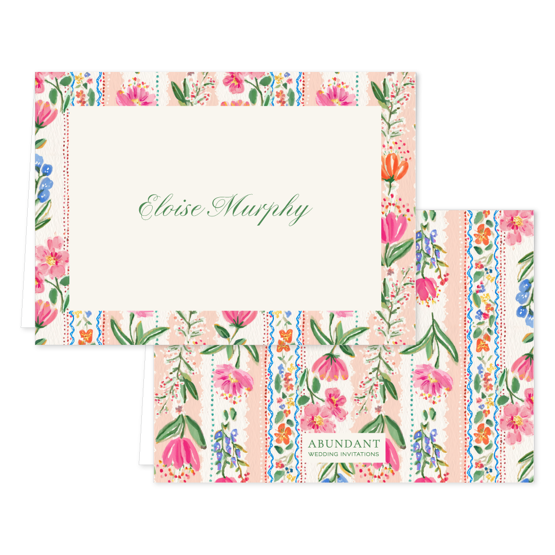 Folded notecard with colorful floral borders and elegant script name on cream background, matching floral envelope behind.