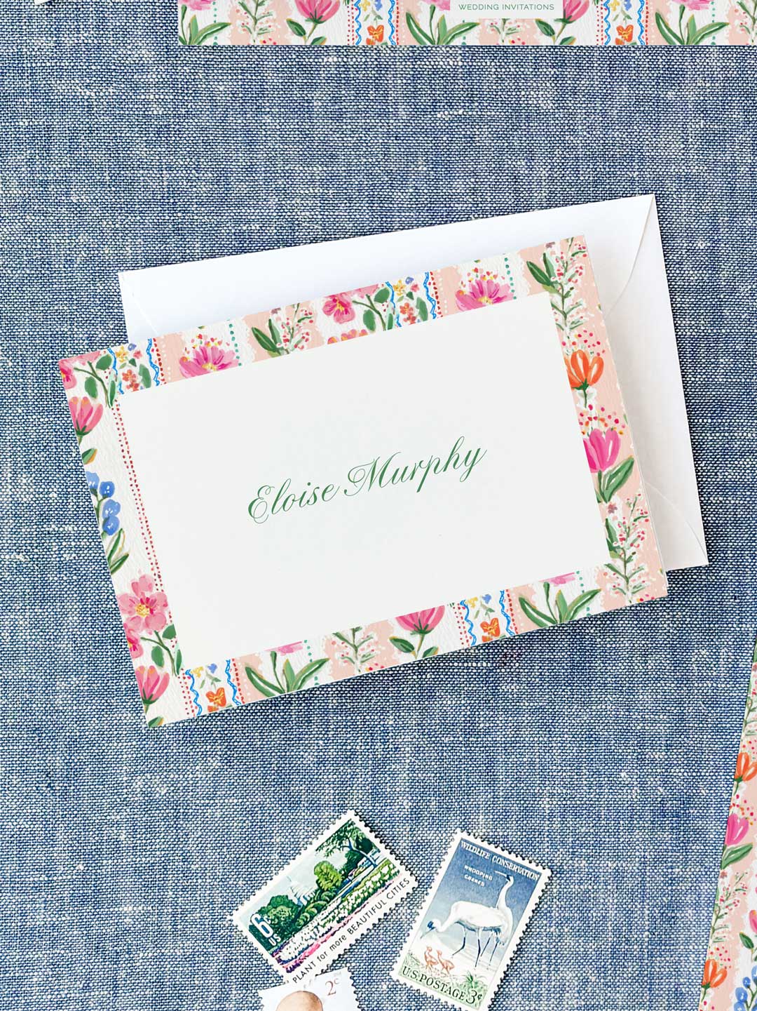 Folded notecard with colorful floral border and elegant green script name on textured blue fabric background.