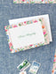 Folded notecard with colorful floral border and elegant green script name on textured blue fabric background.