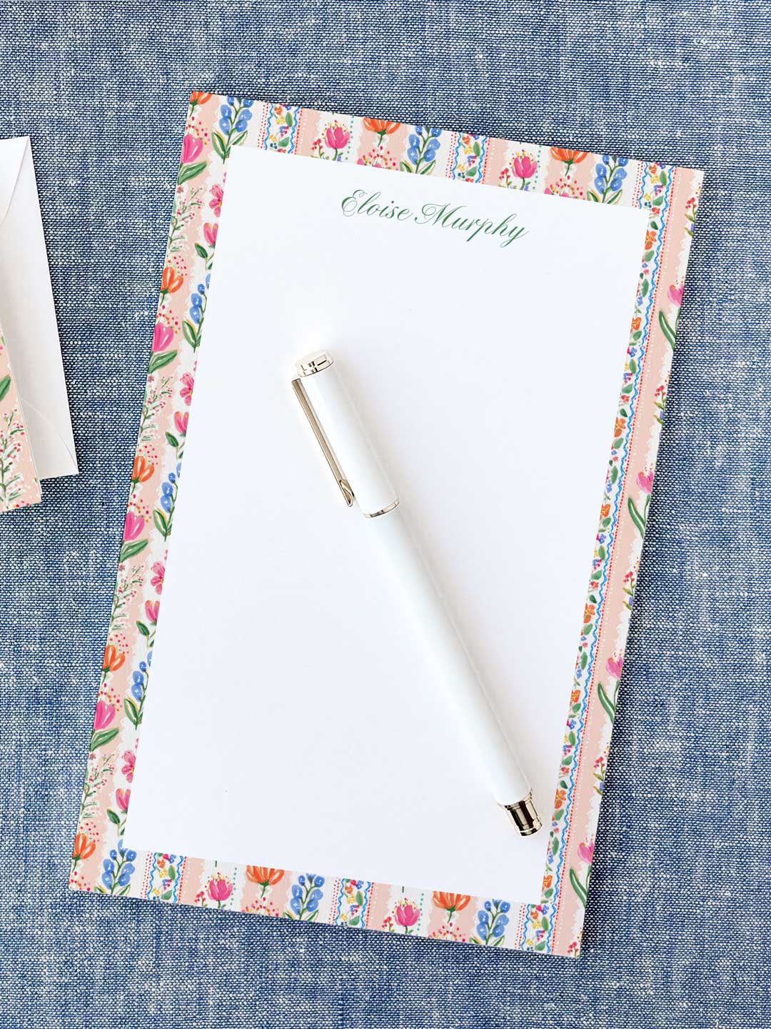 Notepad with colorful floral border and personalized name at the top, next to a white pen on blue fabric.