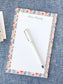 Notepad with colorful floral border and personalized name at the top, next to a white pen on blue fabric.