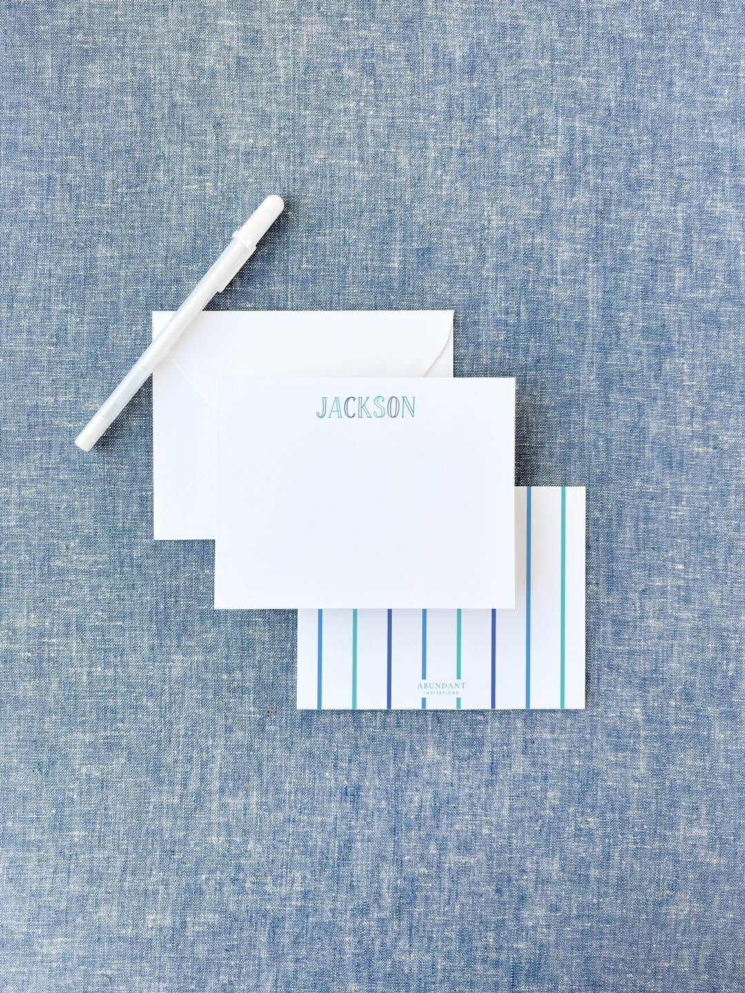 White flat notecard with blue chalkboard-style letters spelling a name, paired with a striped backing card and white envelope on textured blue fabric background.