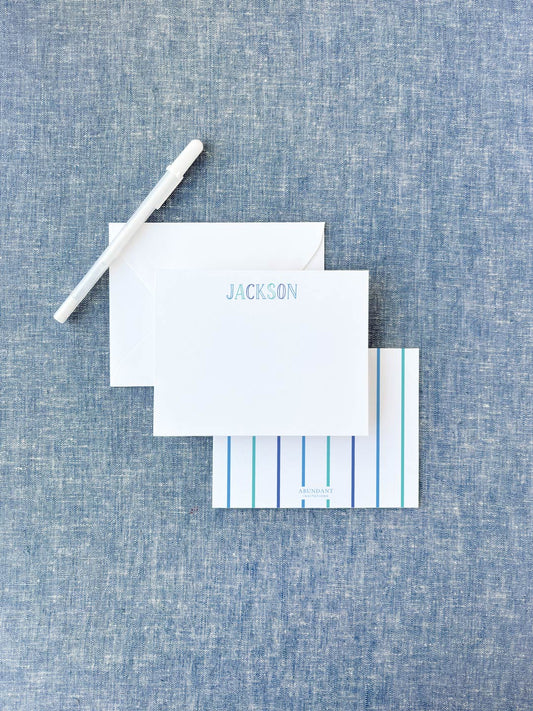 White flat notecard with blue chalkboard-style letters spelling a name, paired with a striped backing card and white envelope on textured blue fabric background.