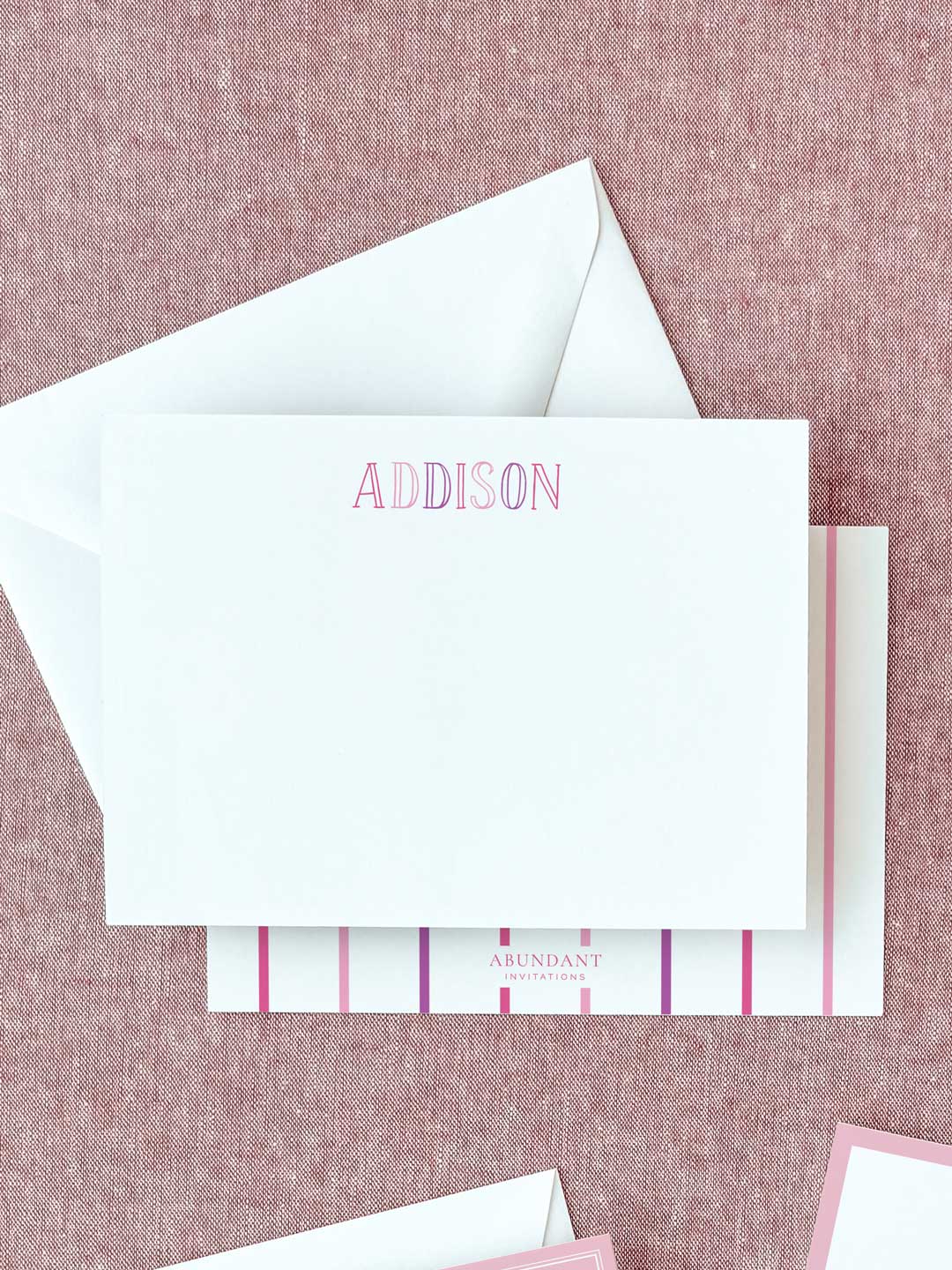 White flat notecard with personalized pink chalkboard-style letters on textured pink fabric background with matching envelope and branding card beneath.