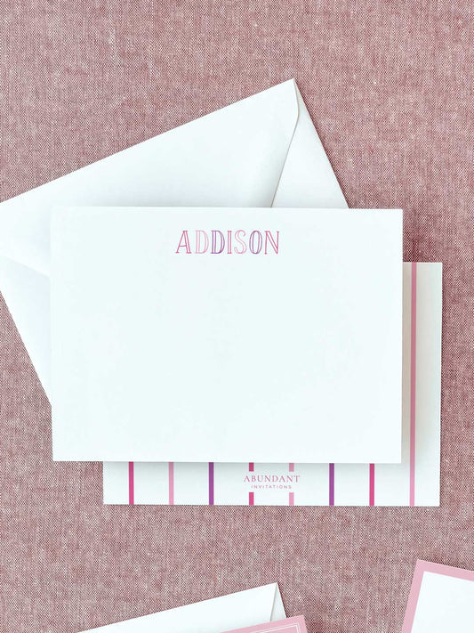 White flat notecard with personalized pink chalkboard-style letters on textured pink fabric background with matching envelope and branding card beneath.