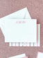 White flat notecard with personalized pink chalkboard-style letters on textured pink fabric background with matching envelope and branding card beneath.