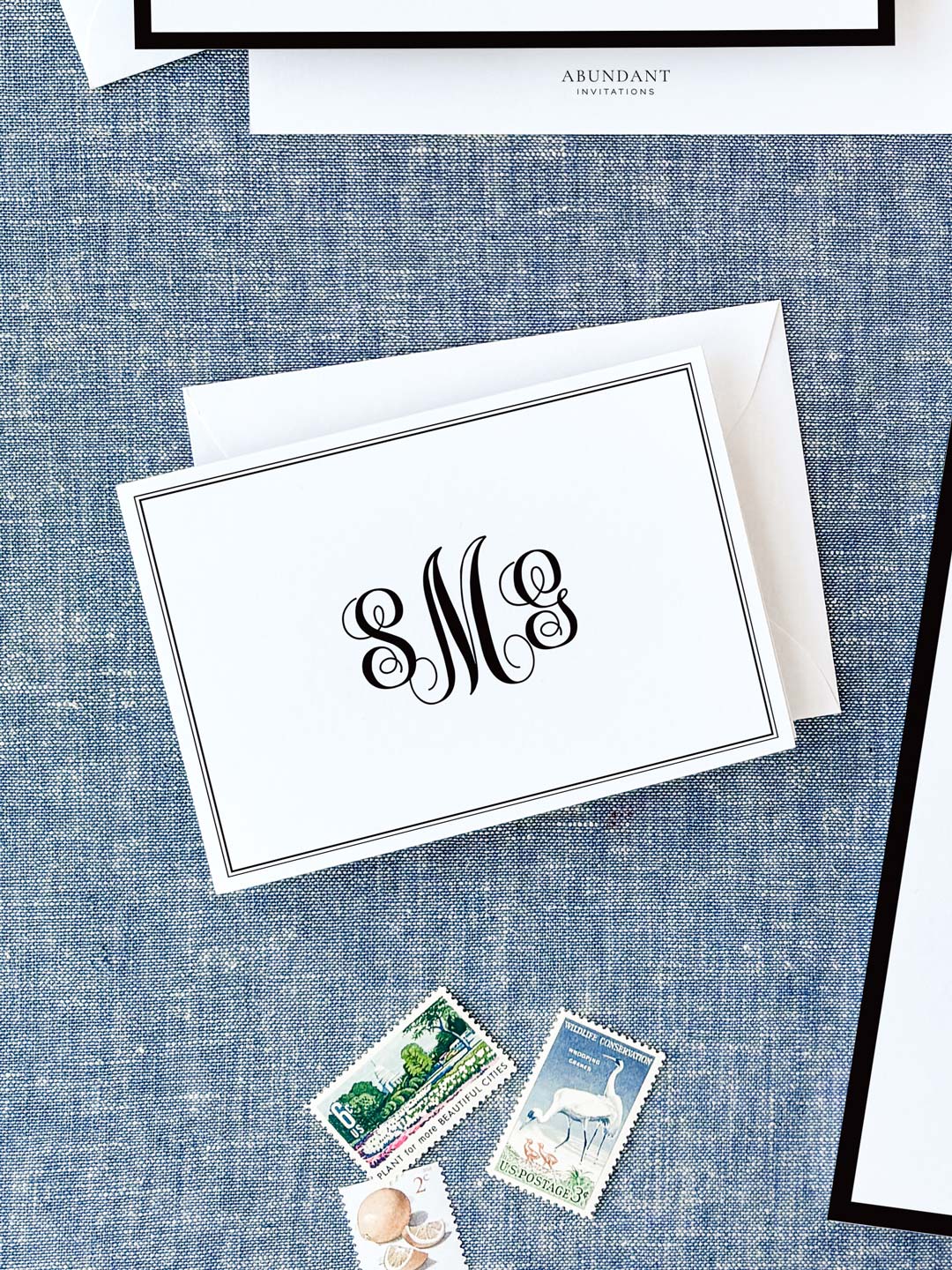 Elegant folded notecard with black monogram initials and a thin double-line border resting on a textured blue fabric background with vintage postage stamps nearby.
