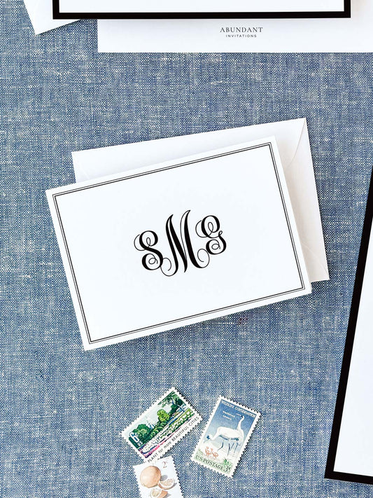 Elegant folded notecard with black monogram initials and a thin double-line border resting on a textured blue fabric background with vintage postage stamps nearby.
