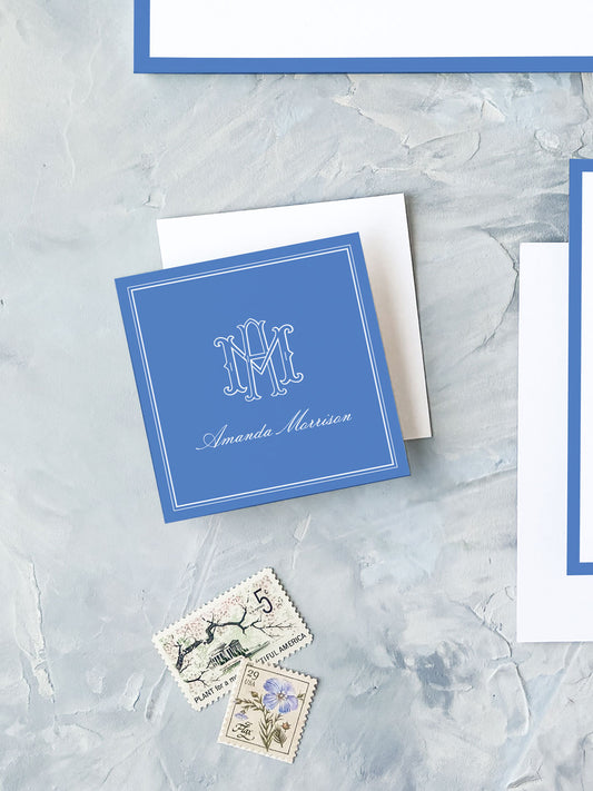 Square cobalt blue personalized calling card with elegant white monogram and cursive name on textured gray background with vintage postage stamps nearby