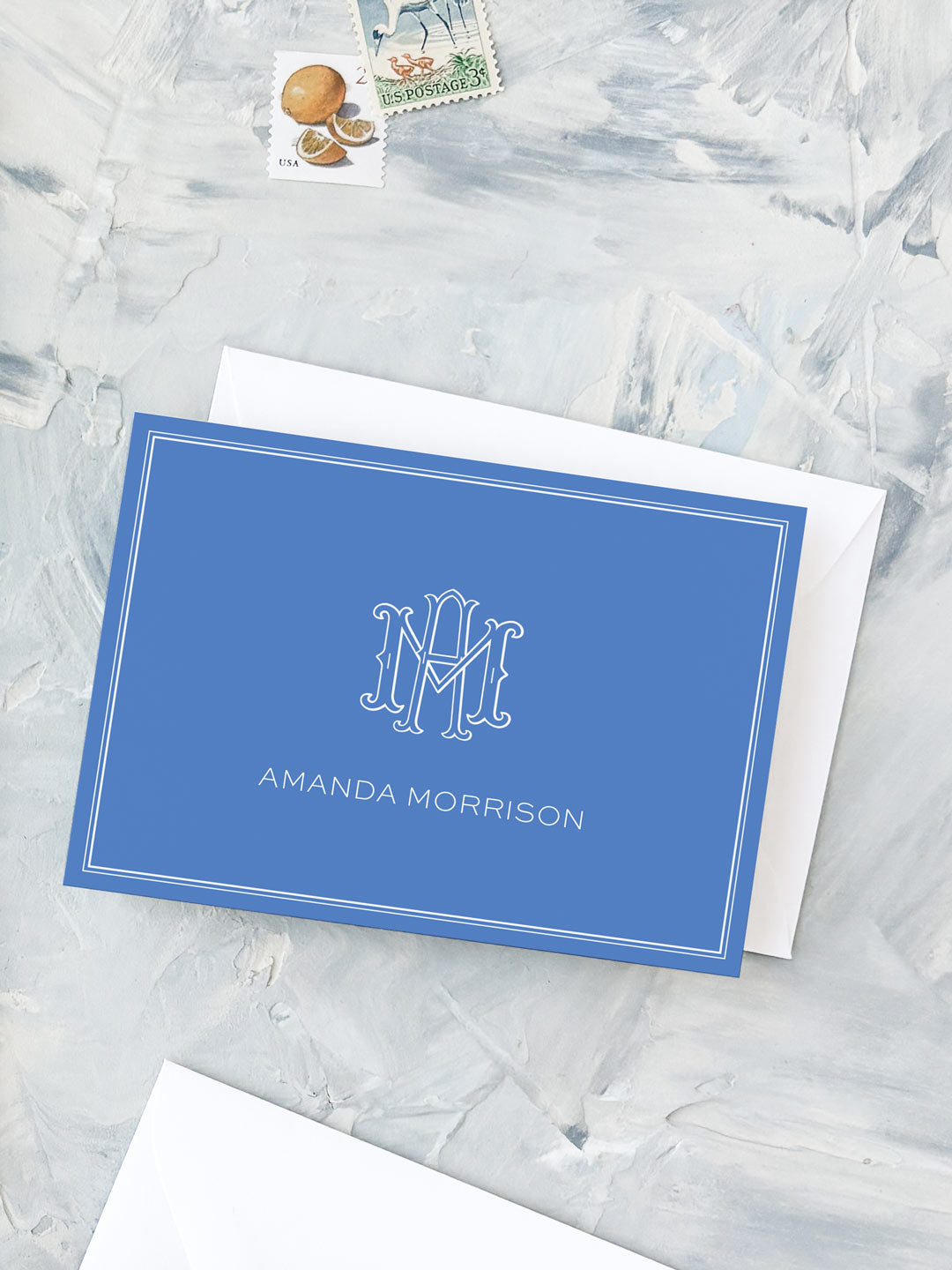Folded cobalt blue notecard with white monogram and personalized name on textured gray surface with postage stamps