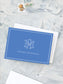 Folded cobalt blue notecard with white monogram and personalized name on textured gray surface with postage stamps