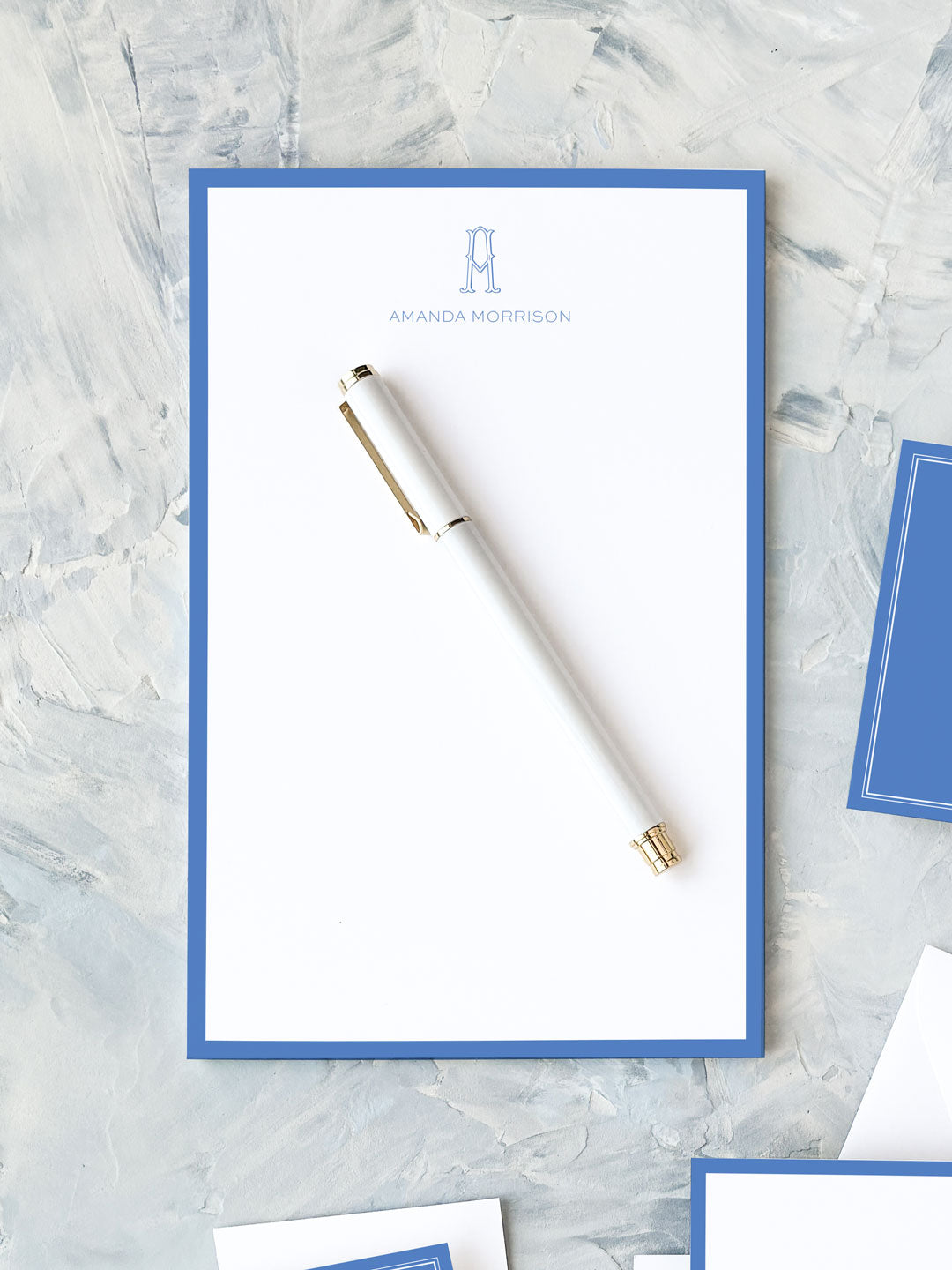 Elegant notepad with a classic cobalt border and personalized monogram, styled with a sleek white pen on textured surface.