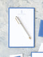 Elegant notepad with a classic cobalt border and personalized monogram, styled with a sleek white pen on textured surface.