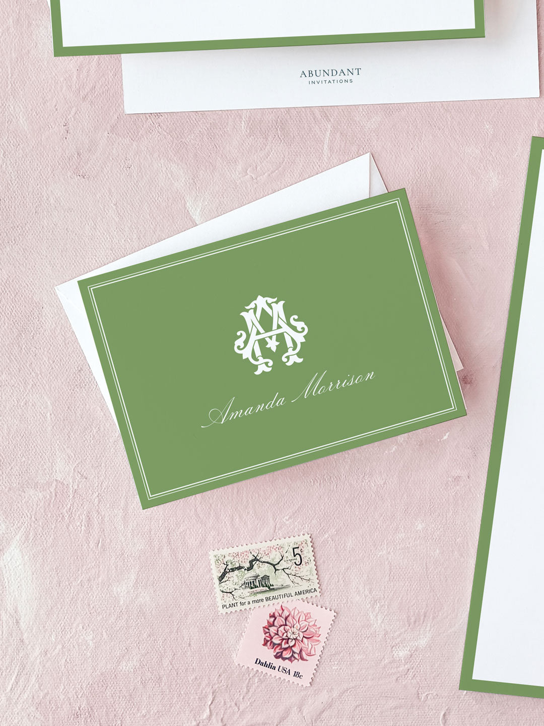 Elegant green folded notecard featuring a classic monogram and personalized script, displayed with vintage postage on a soft pink background.