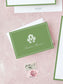Elegant green folded notecard featuring a classic monogram and personalized script, displayed with vintage postage on a soft pink background.