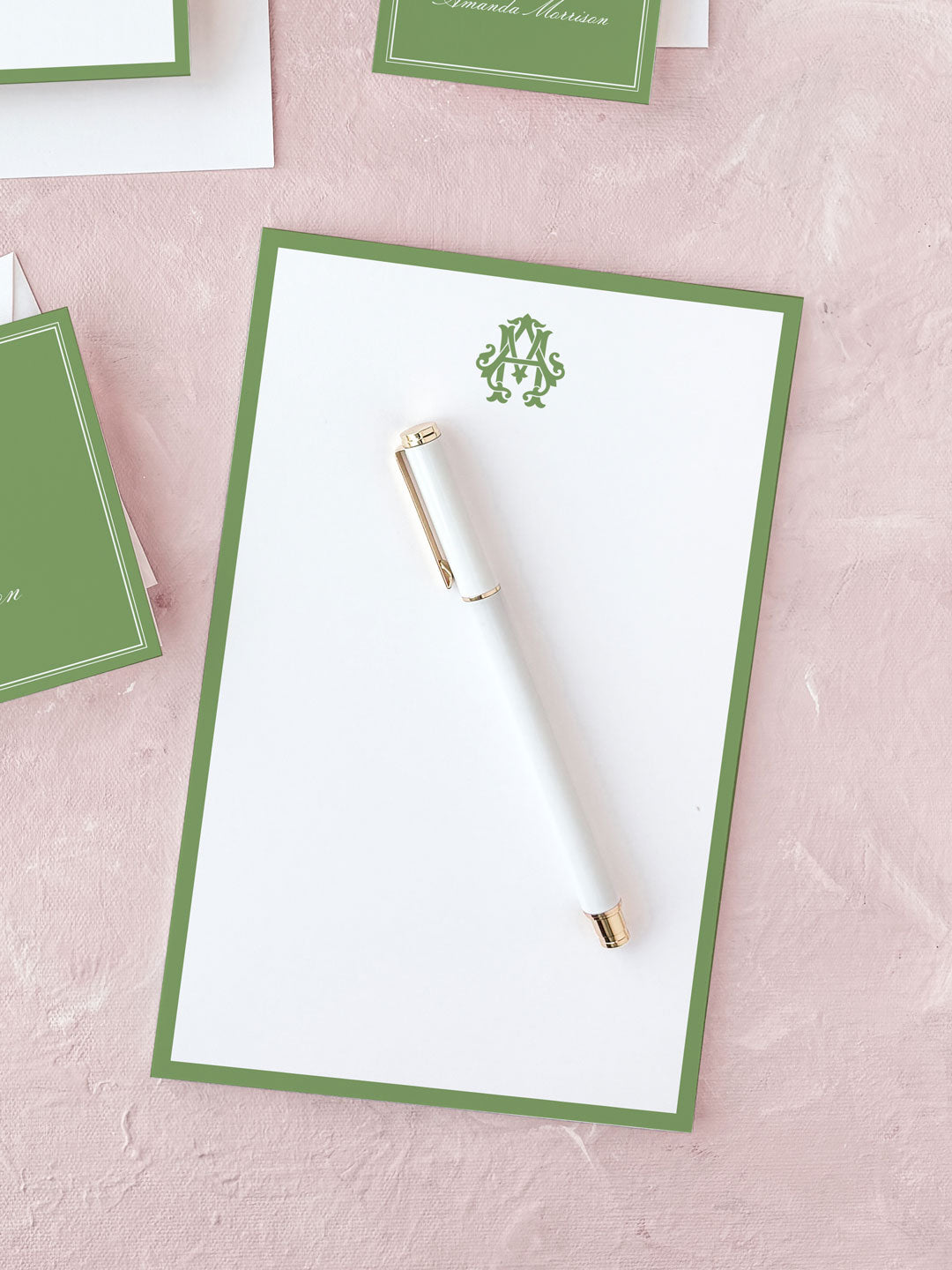 White notepad with a green border and monogram at the top, accompanied by a white pen with gold accents on a pink textured surface.
