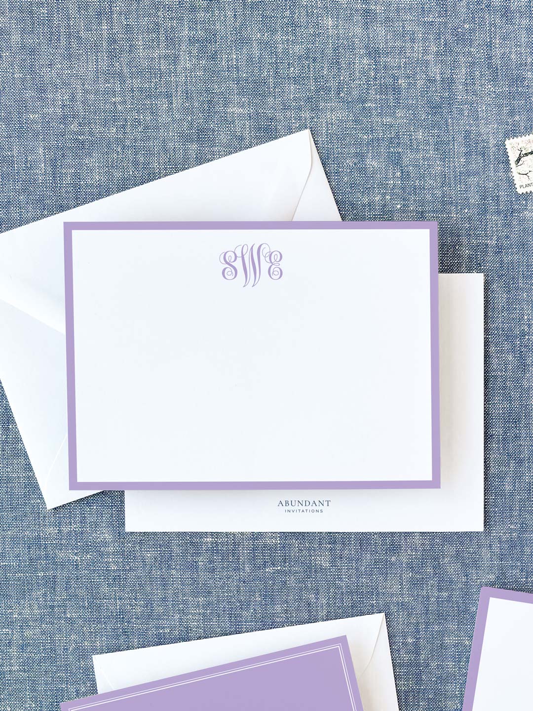 Elegant white flat notecard with a lavender border and monogram, paired with matching envelope on textured fabric background.