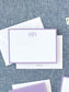 Elegant white flat notecard with a lavender border and monogram, paired with matching envelope on textured fabric background.