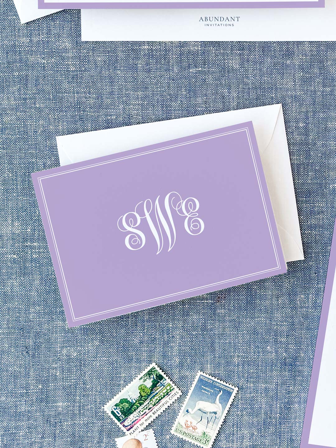Folded lavender notecard with elegant white monogram on textured blue background, paired with white envelope and vintage postage stamps.