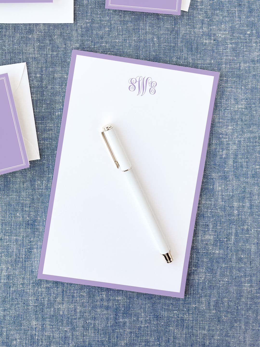 Notepad with a soft lavender border and elegant monogram lettering at the top, accompanied by a sleek white pen on a textured blue surface.