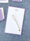 Notepad with a soft lavender border and elegant monogram lettering at the top, accompanied by a sleek white pen on a textured blue surface.